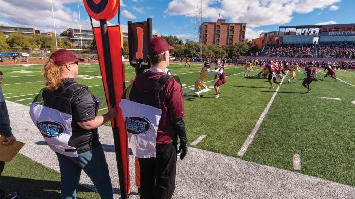 Dennis Ugolini and Jennifer Steele watch a Trinity Football game from the sidelines with the down and distance markers